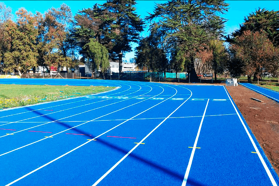 The blue athletics track at Temuco looking along the straight into the bend, white lane lines and coloured relay marks on the surface, mature trees and university buildings beyond.