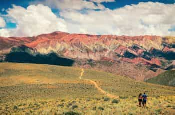 A couple walks up a hill covered in green and yellow shrubs at the base of red mountains in Jujuy, Argentina.