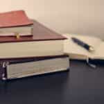 Three heavy books stacked on top one another on a desk next to an open notebook with a pen on top.